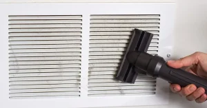 Hand vacuuming dust from a dirty air vent grille during $99 air duct cleaning.